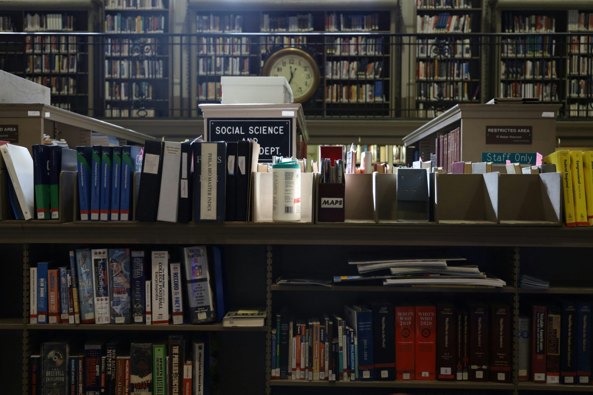"A well-lit library scene featuring neatly organized shelves filled with books and binders in the Social Science department. A vintage clock is visible in the background, adding a timeless touch to the serene academic atmosphere. The image captures the essence of a quiet, knowledge-filled space."